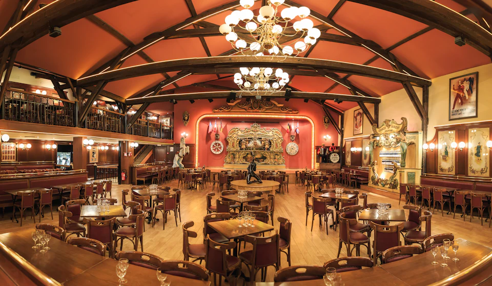 Large, ornate dining hall with wooden tables and chairs, chandeliers, stage with elaborate decor, and a mezzanine level.