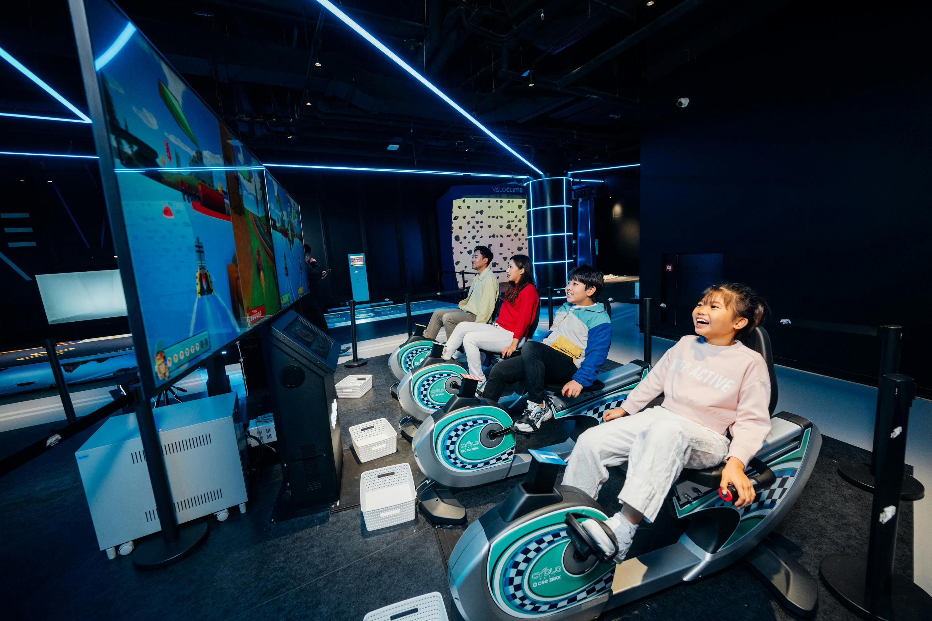 Children sitting on interactive ride simulators in a futuristic indoor entertainment space with neon lighting.