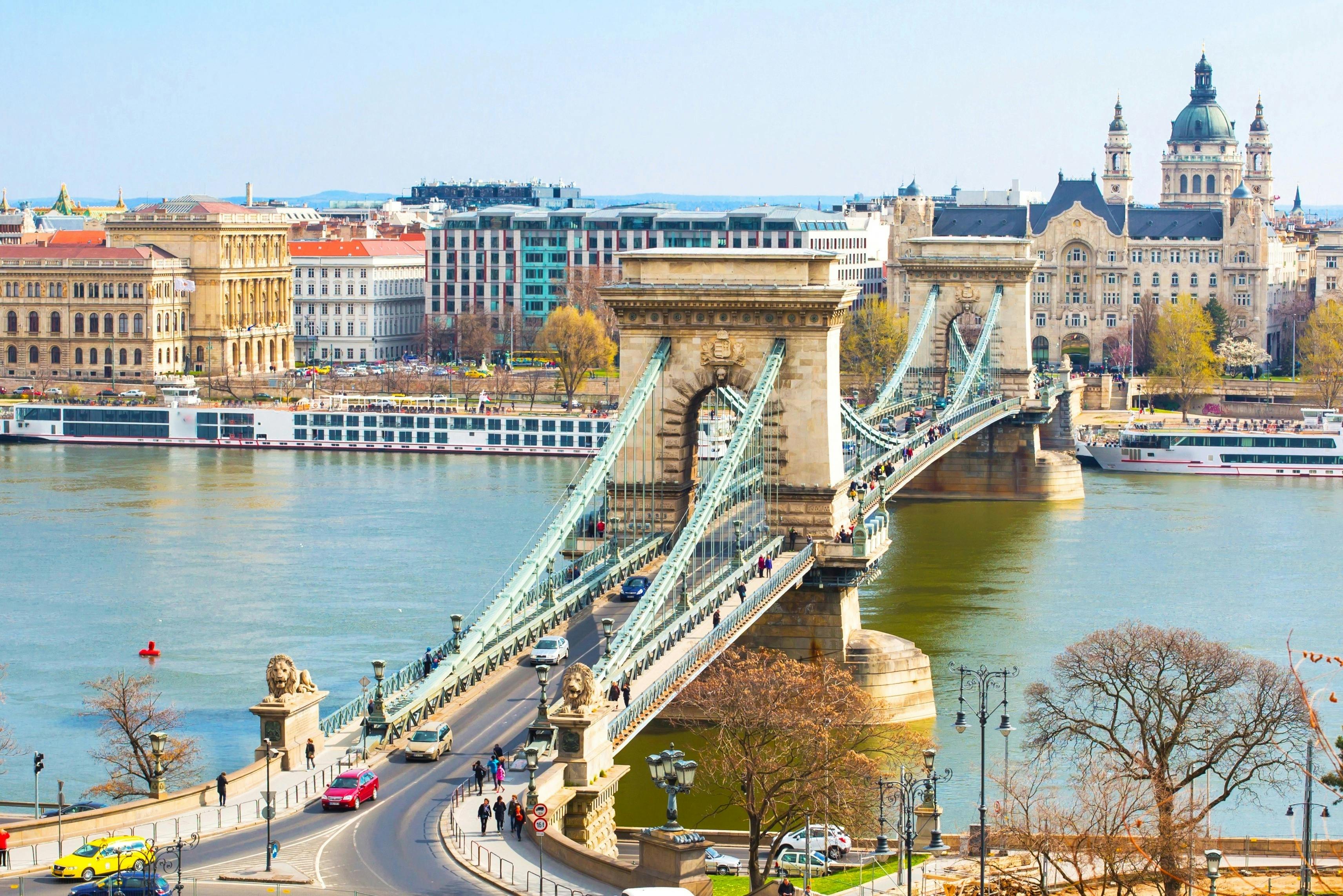 A suspension bridge with stone towers spans a river in a cityscape, featuring vehicles, pedestrians, and surrounding buildings.