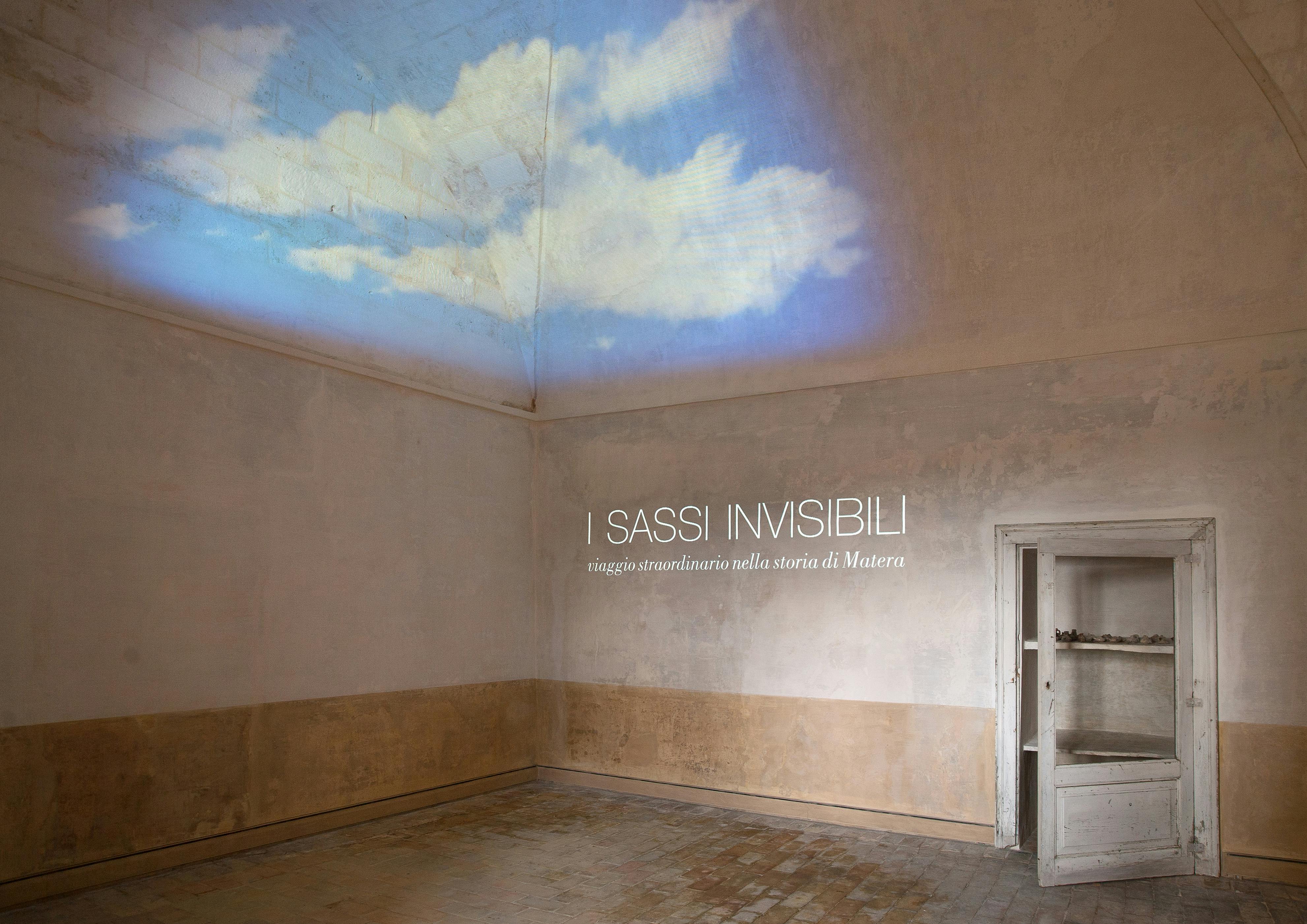 Rustikální, prázdná místnost s otevřenými dveřmi a projekcí mraků na stropě. Text zní: „I Sassi Invisibili: viaggio straordinario nella storia di Matera.“