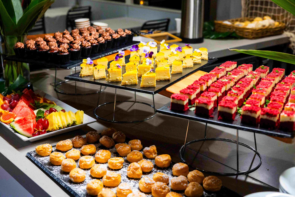 An assortment of desserts on trays, including chocolate cupcakes, yellow cakes with flowers, red-topped bars, and powdered donut holes.