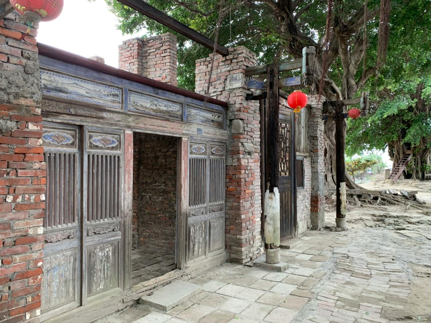 An old brick and wood building with wooden doors and red lanterns hanging, surrounded by trees on a paved pathway.