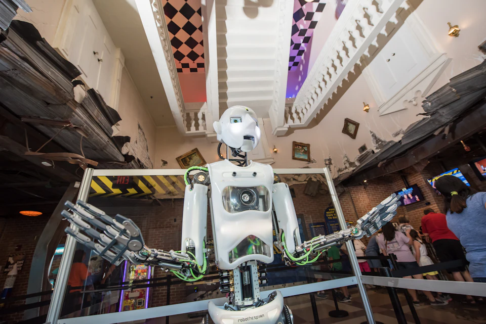 A humanoid robot with exposed mechanics stands in a whimsical, museum-like setting with tilted picture frames and a checkered ceiling.