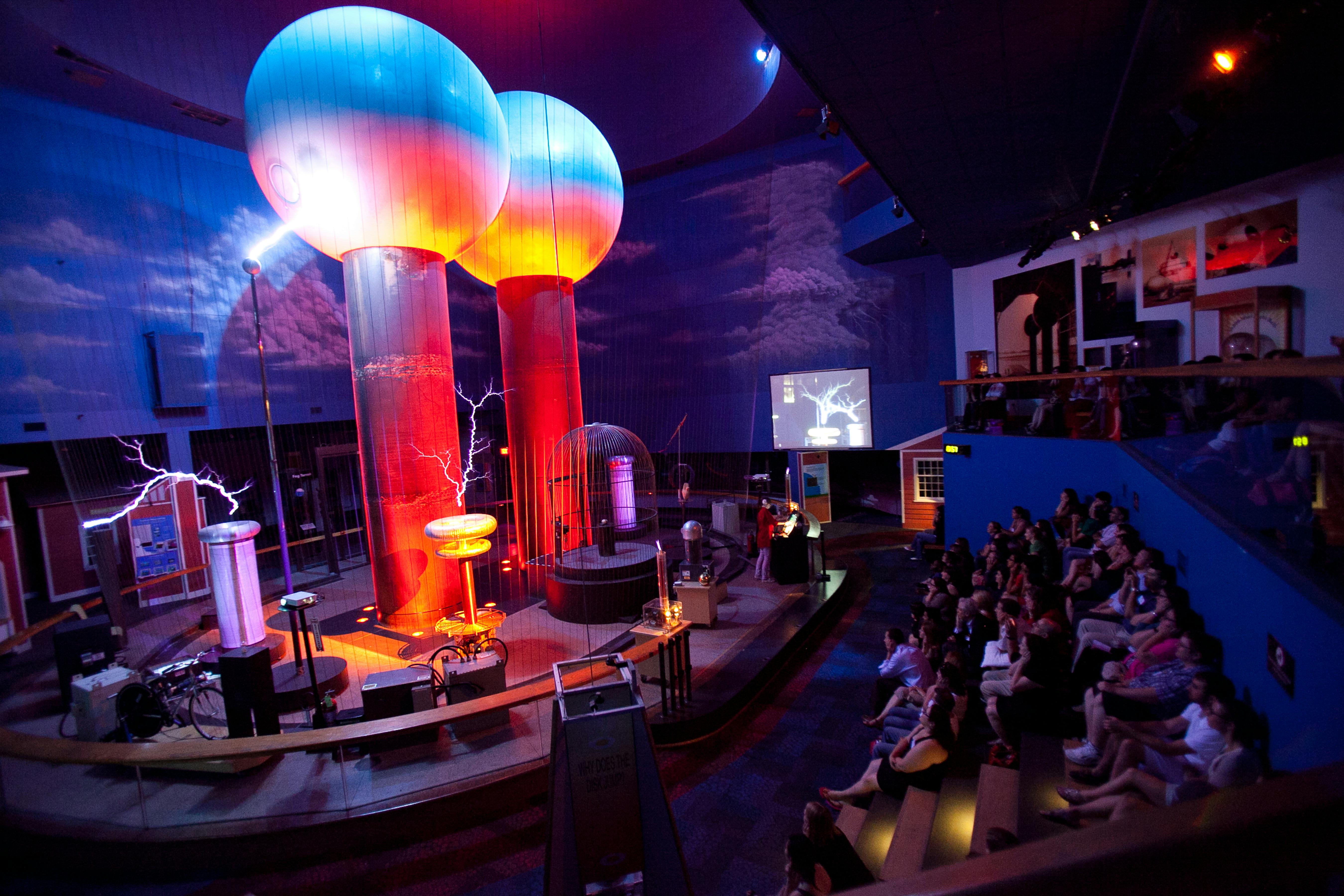 관중들은 강당에서 대형 테슬라 코일과 전기 디스플레이가 있는 생동감 넘치는 과학 시연을 지켜보고 있습니다.