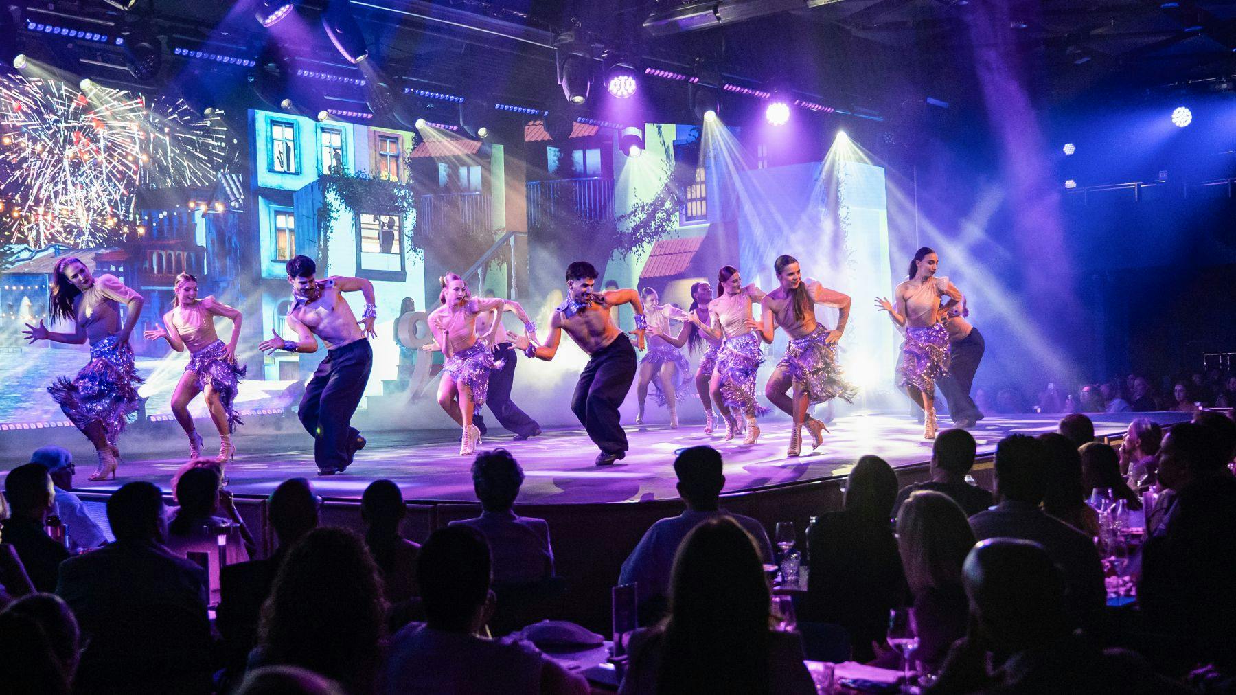 Um grupo de bailarinos actua num palco bem iluminado com um pano de fundo colorido, visto por um público sentado em mesas.