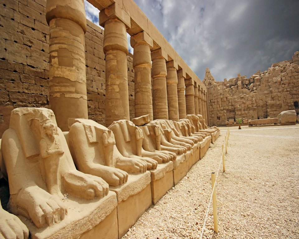 Row of ancient stone sphinx statues with lion bodies and ram heads beside large columns in a historic ruin under a cloudy sky.