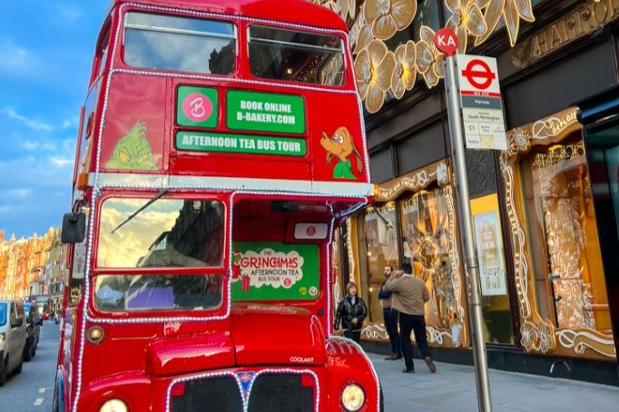 A red double-decker bus promoting an afternoon tea tour is parked near a decorated building and a bus stop in an urban area.