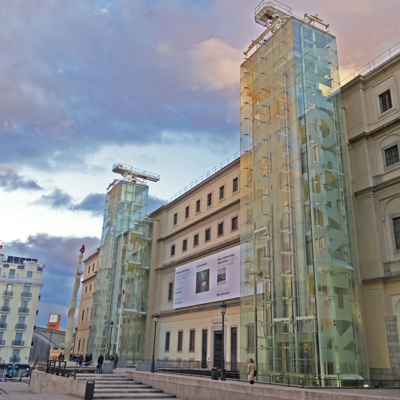 En modern byggnad med glashissar märkta "REINA SOFIA" och affisch, mot en molnig himmel, människor som går i förgrunden.