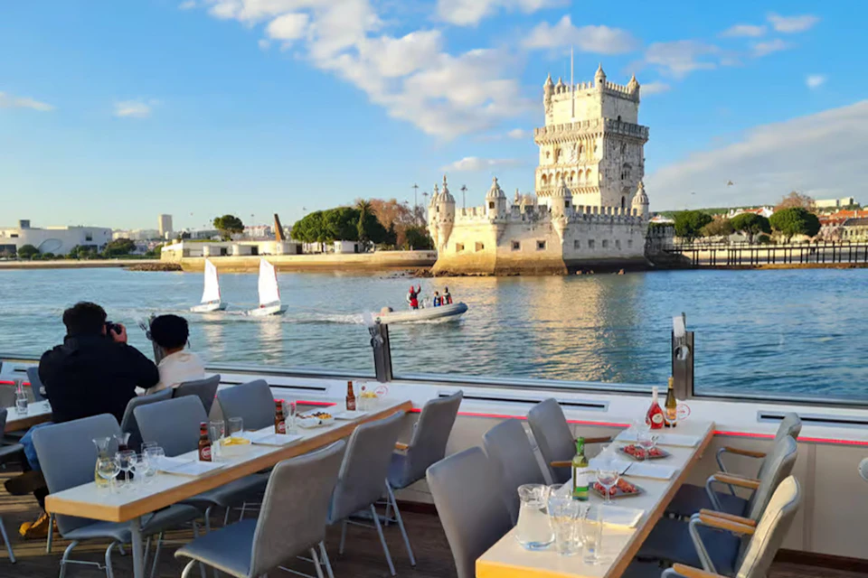 Outdoor dining area on a boat with a view of the Belem Tower, sailboats, and a motorboat on a sunny day.