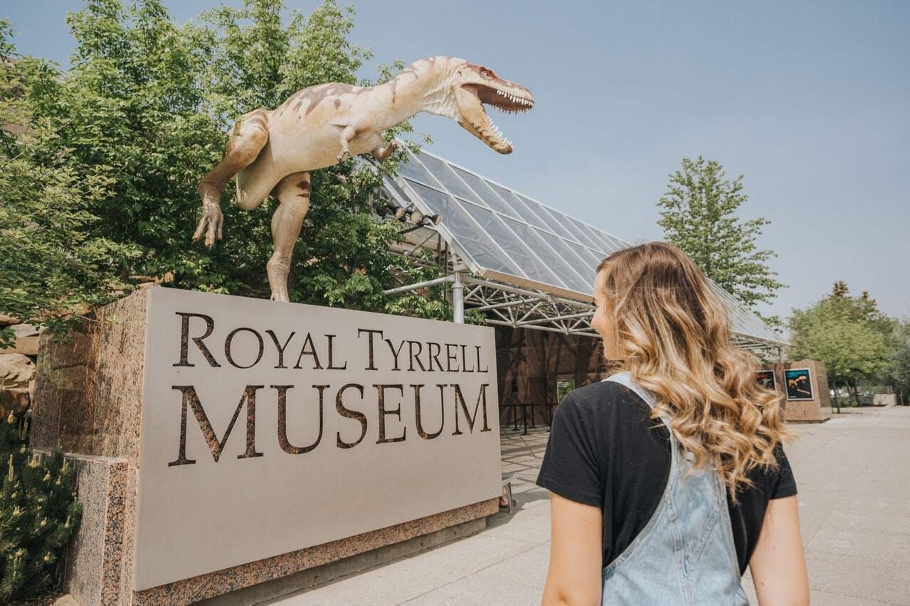 Una persona si trova vicino a un cartello con la scritta &#34;Royal Tyrrell Museum&#34;, con una statua di dinosauro e alberi sullo sfondo.
