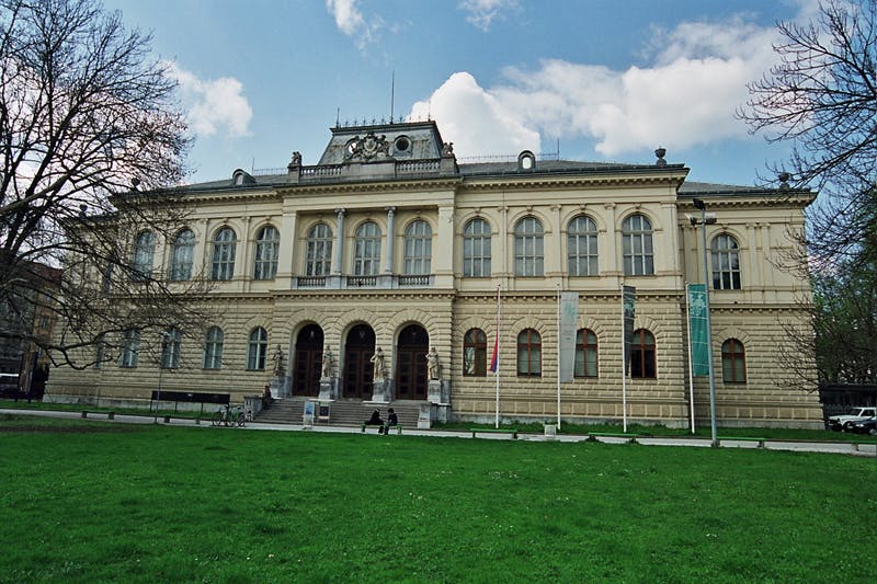 Historická béžová budova s klenutými okny a prapory na sloupech, zasazená za zelený trávník a pod částečně zataženou oblohu.