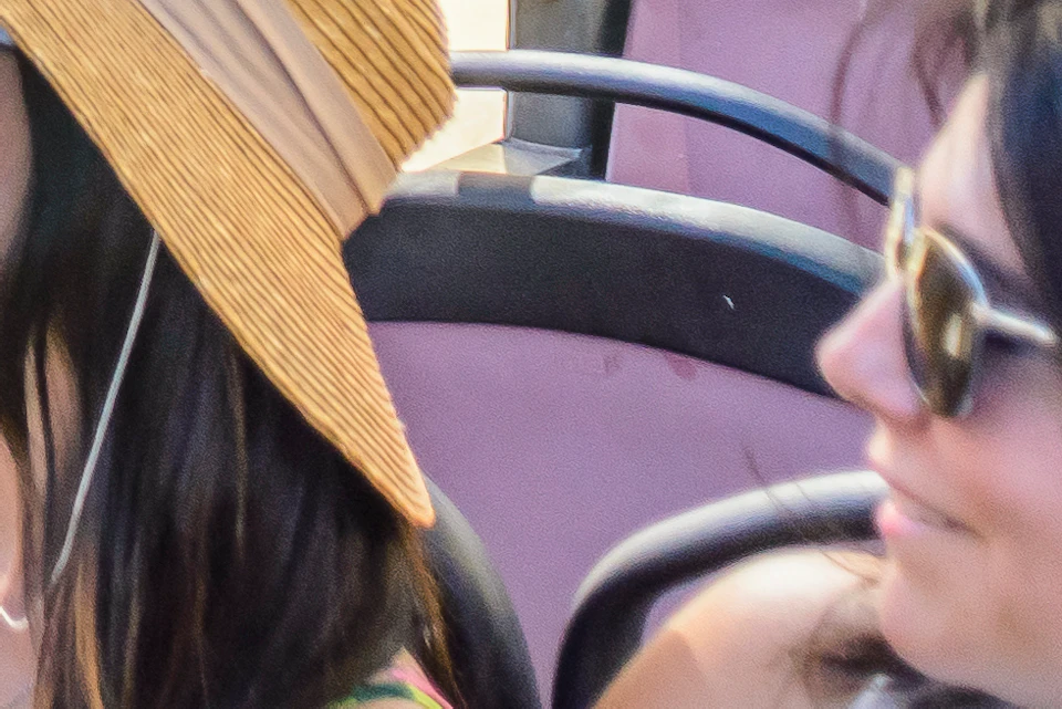 Two women seated on the top deck of an open-top bus, one taking a photo. Background shows a large, ornate yellow building with balconies.