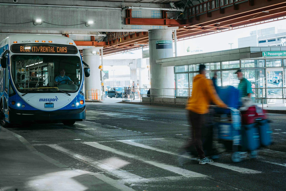 A person in casual attire with a hat and camera walks past a shuttle bus labeled "ORIO SHUTTLE."