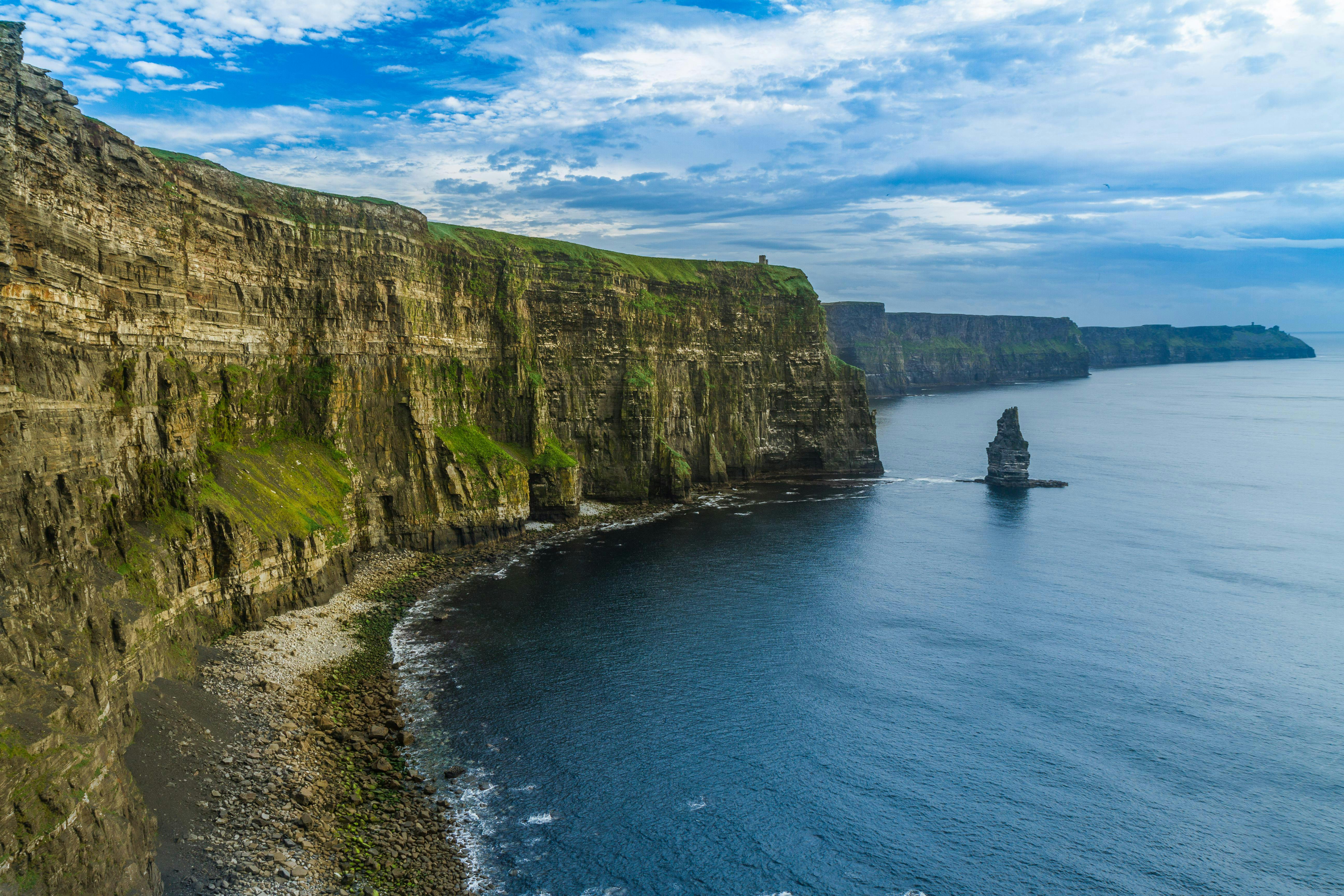 Steep sea cliffs covered in green vegetation, with rocky shores and calm blue waters under a partly cloudy sky.