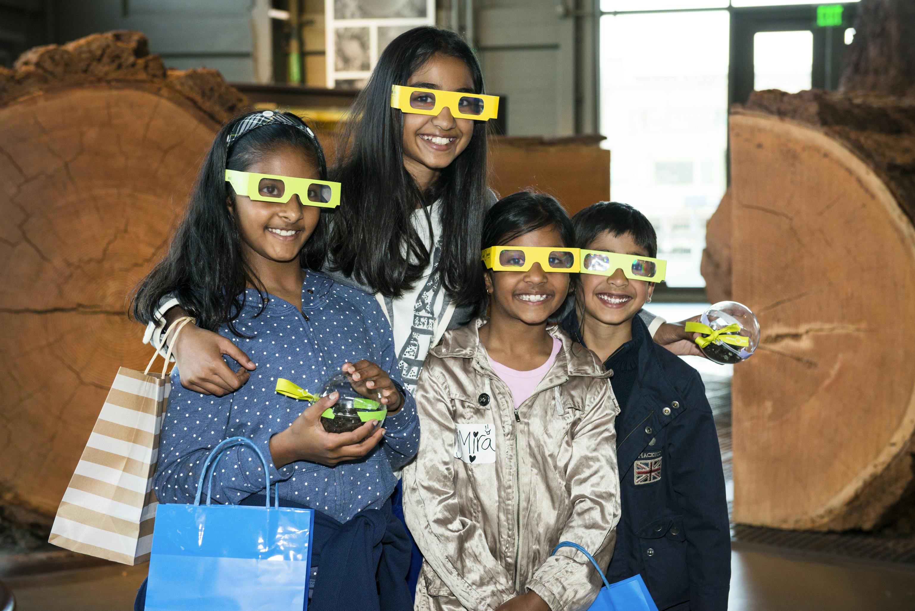 Four smiling children wearing 3D glasses hold blue gift bags and small objects, standing indoors with wooden structures in the background.