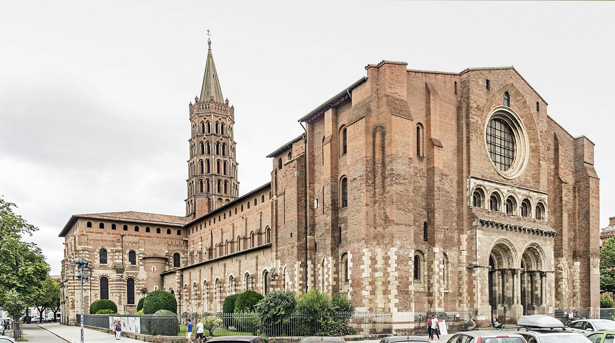 Historic brick church with a tall, detailed bell tower, arcaded entrance, and large circular window; a few people and cars visible.