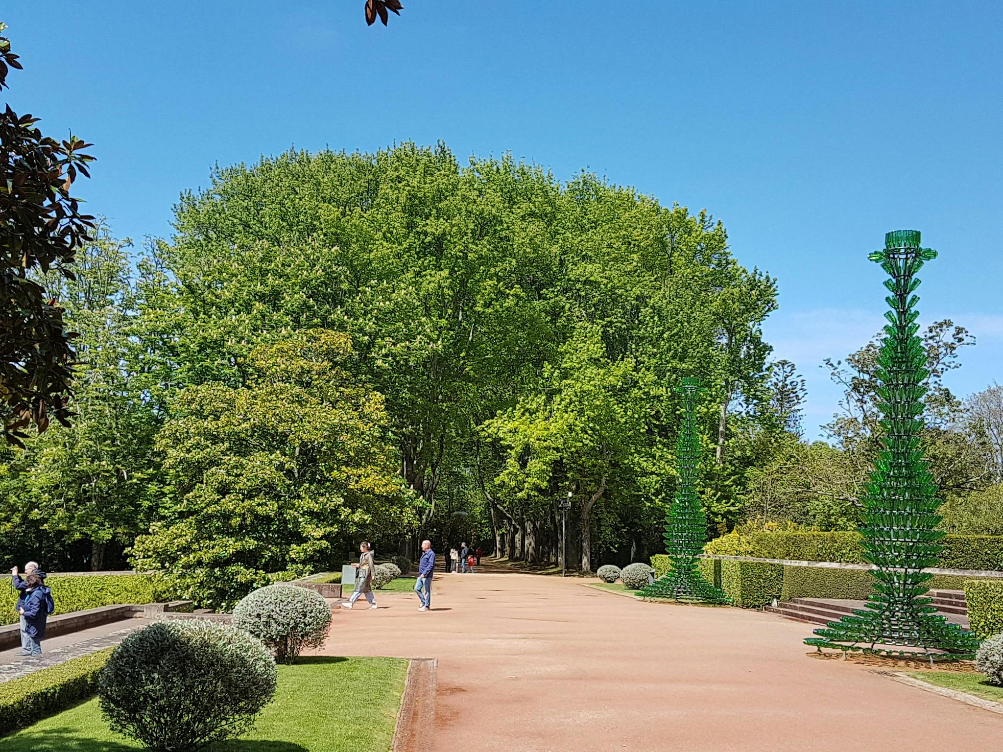 En rymlig gångväg i parken flankerad av trimmade buskar, träd och tre personer som går under en klarblå himmel. Två gröna skulpturer är närvarande.