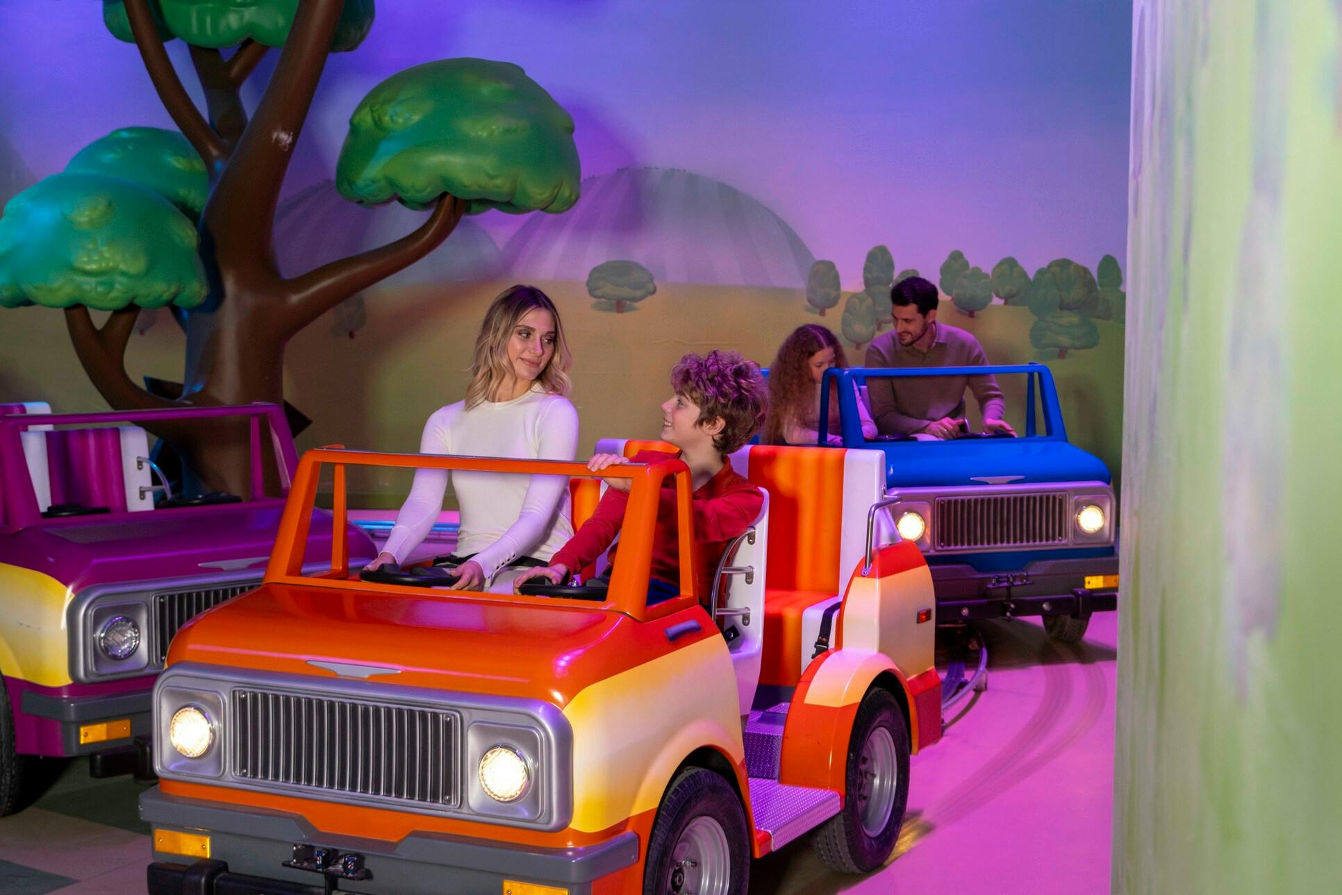 Mensen rijden op kleine, kleurrijke voertuigen in een amusementsthema met cartoons, met geschilderde bomen en heuvels op de achtergrond.