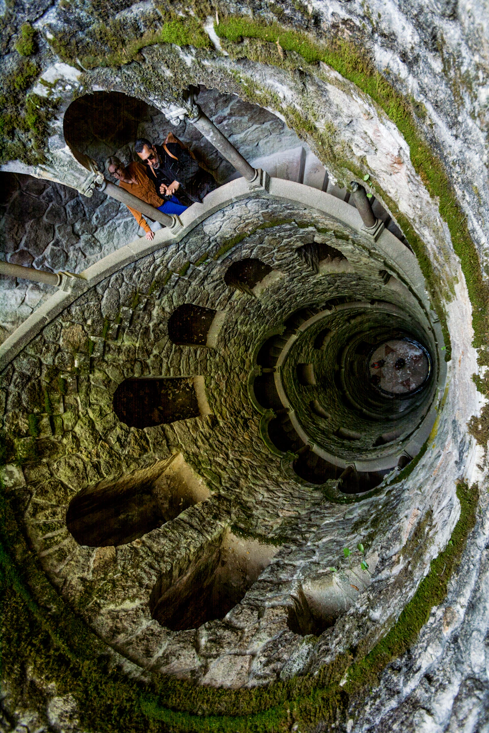 A person in a brown jacket looks down into a stone spiral staircase with multiple arched openings and moss-covered walls.