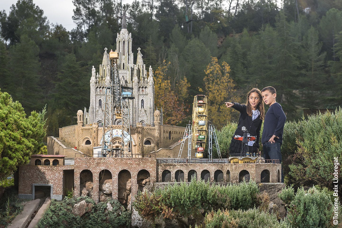 Two children observe and point at a detailed miniature model of a historic building and Ferris wheel amidst lush greenery.