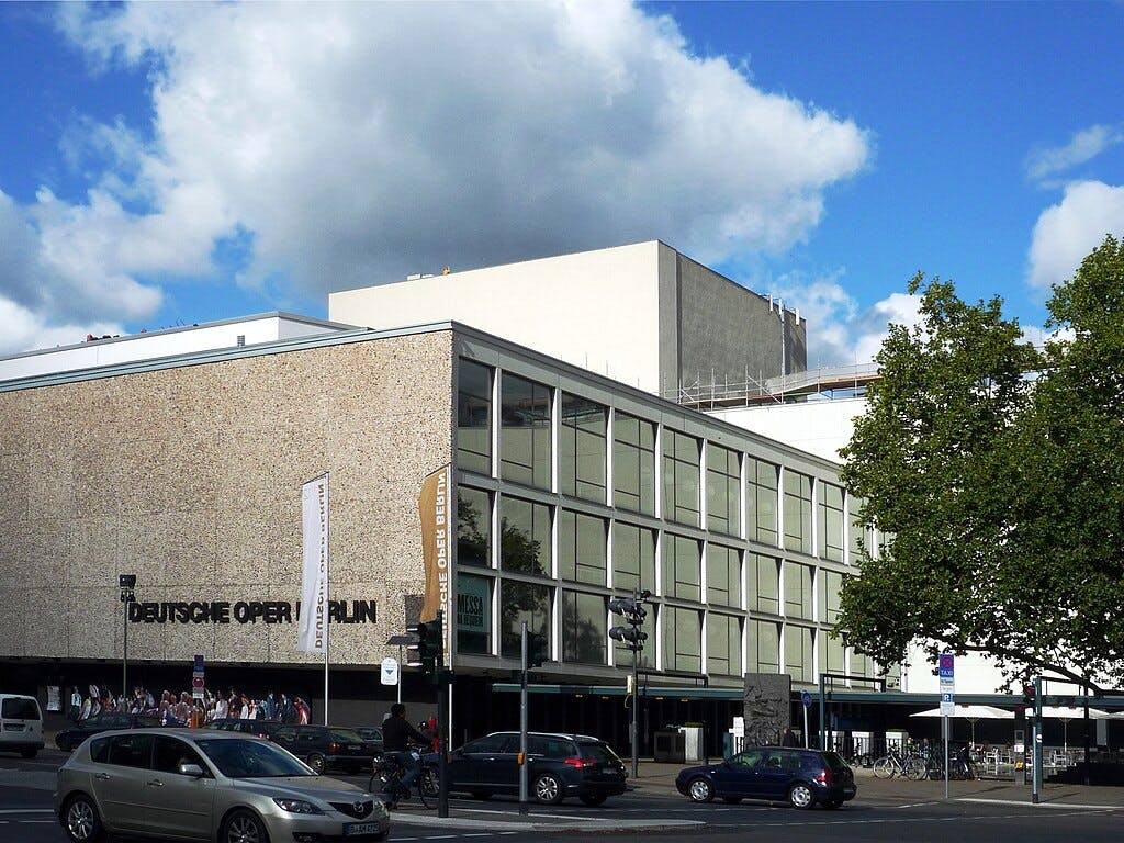 「ドイツ・オペラ・ベルリン」と書かれた大きな窓があるモダンな建物。 前方に車と歩行者がいます。木が部分的に見えます。