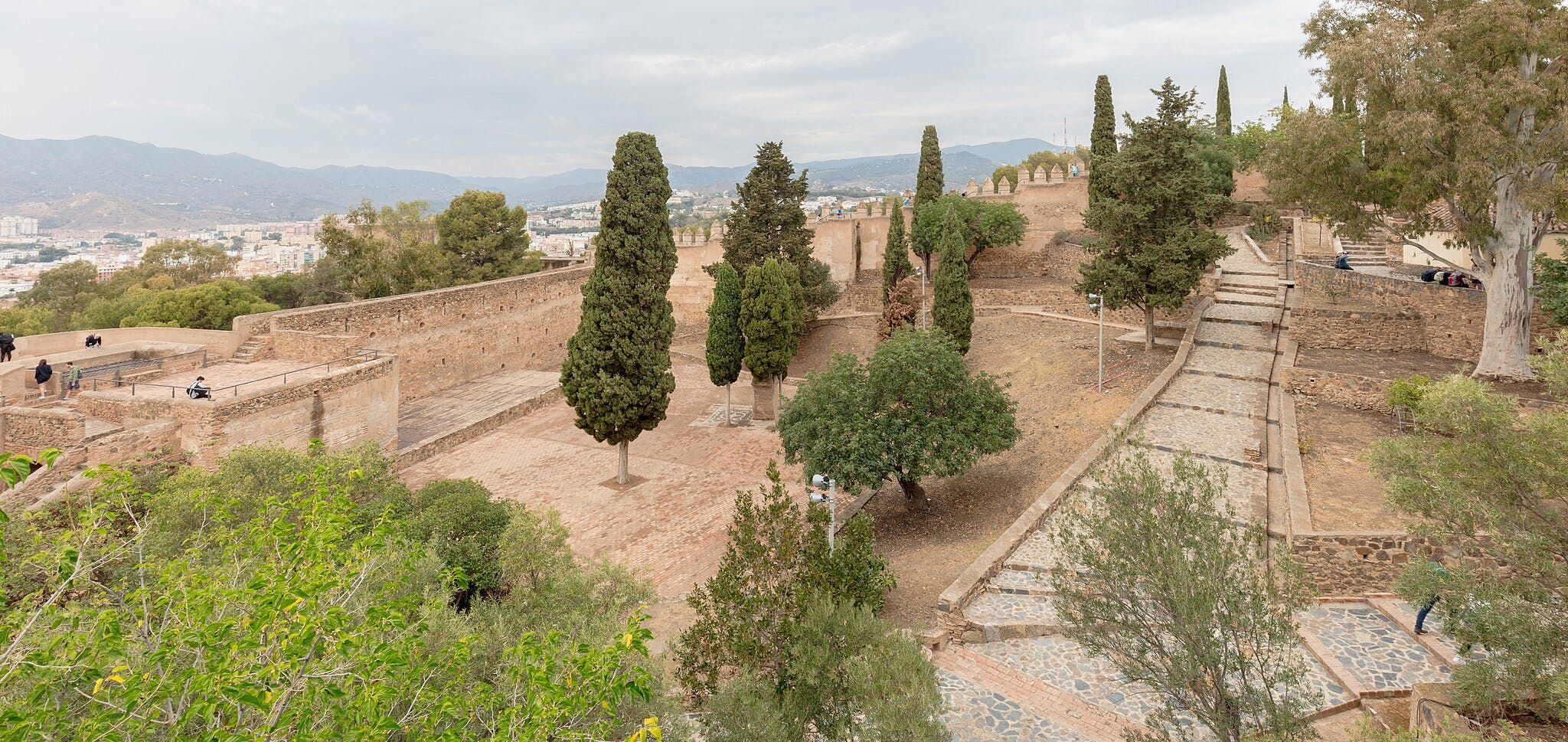 Sito storico con camminamenti in pietra, alberi ad alto fusto e visitatori sparsi. Colline ed edifici cittadini sullo sfondo.