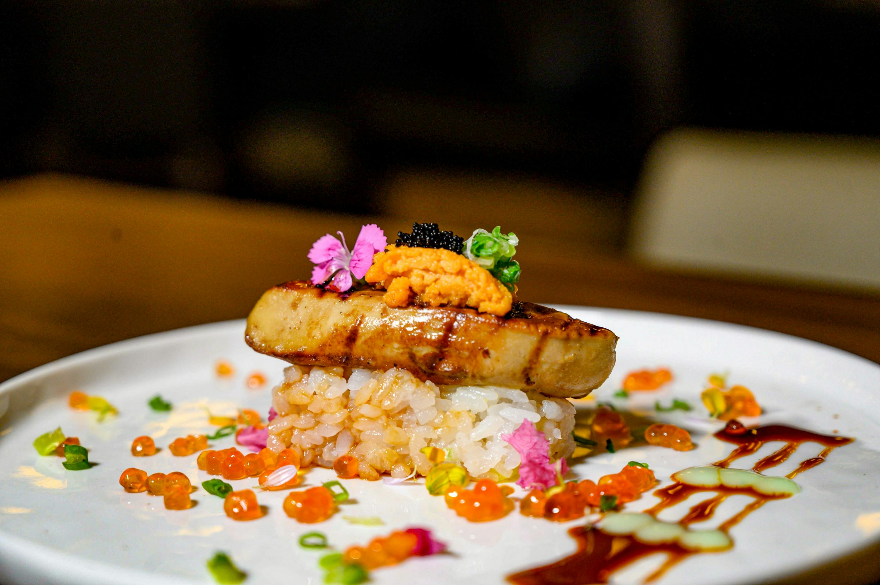A plate of gourmet sushi topped with seared foie gras, uni, caviar, and garnished with roe, flowers, and sauces.