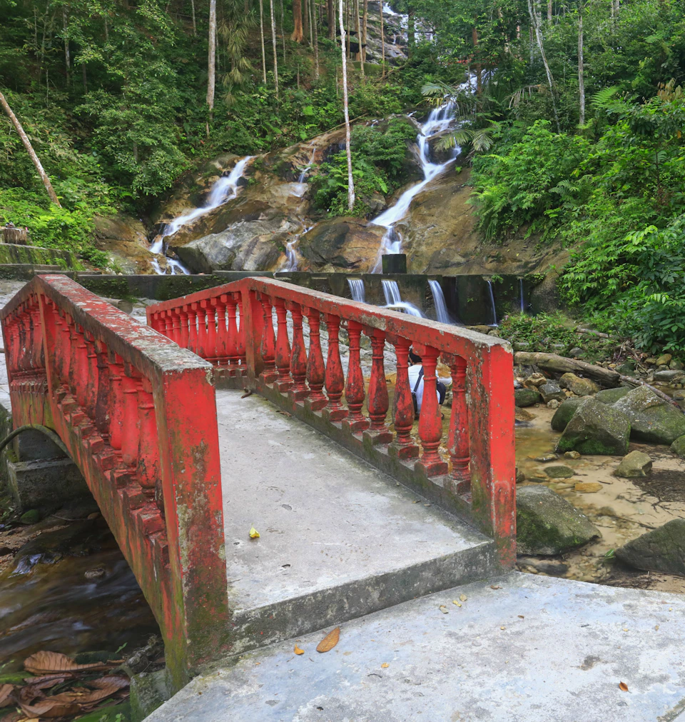 Red bridge over a rocky stream in a lush, green forest with a multi-tiered waterfall in the background.