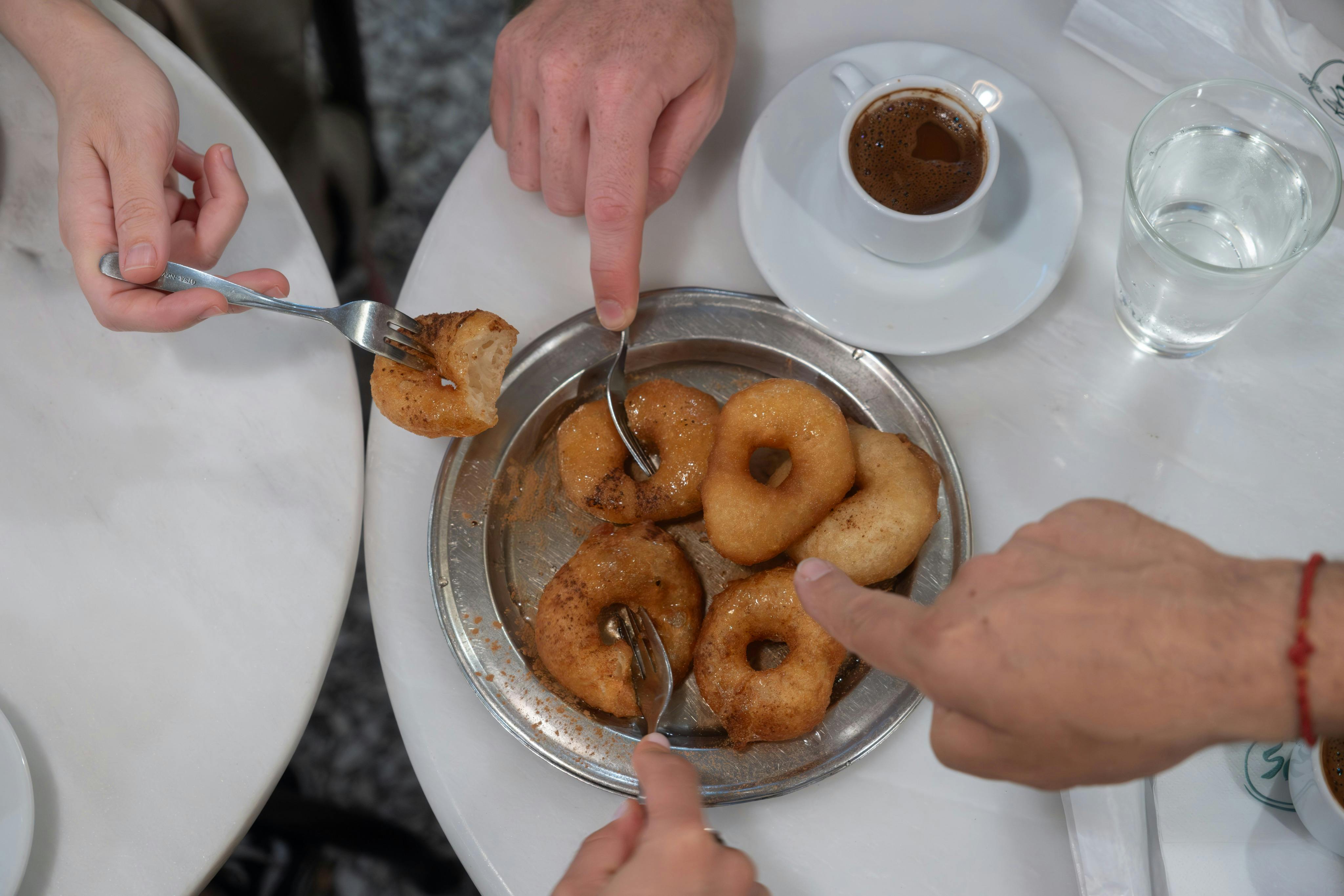 Τρία χέρια φτάνουν για ντόνατς σε ένα ασημένιο πιάτο, με ένα φλιτζάνι καφέ κοντά σε ένα λευκό μαρμάρινο τραπέζι.