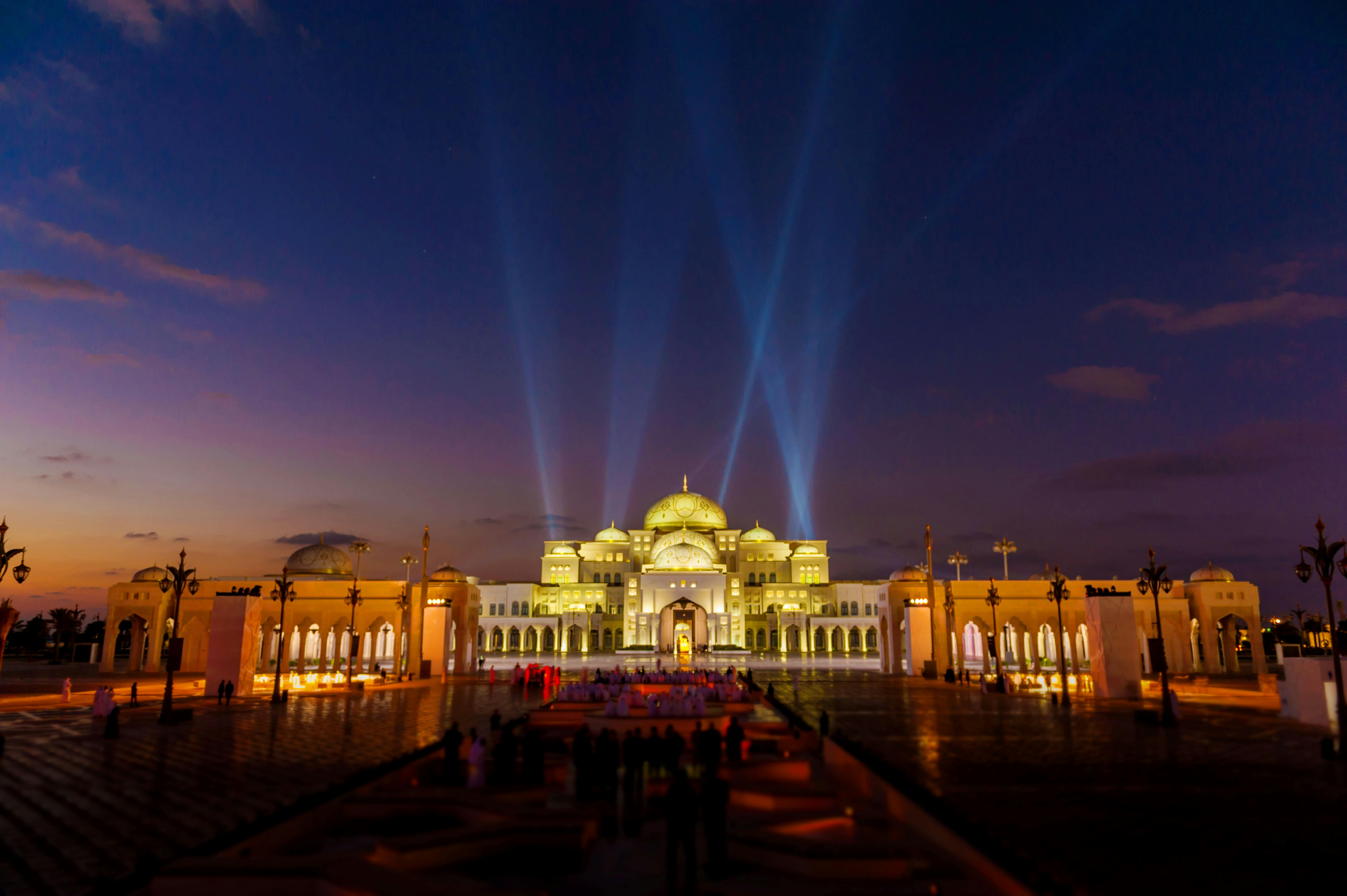 Illuminated palace with domes at dusk, spotlights beaming upwards, reflections on wet ground, and people gathered in the foreground.