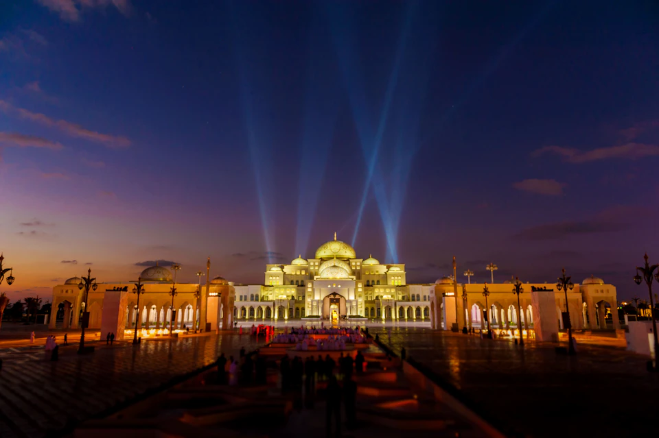 Illuminated palace with domes at dusk, spotlights beaming upwards, reflections on wet ground, and people gathered in the foreground.