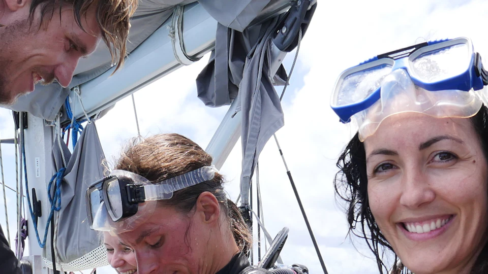 A woman smiles at the camera while two other individuals prepare for scuba diving on a boat, assisted by a man.