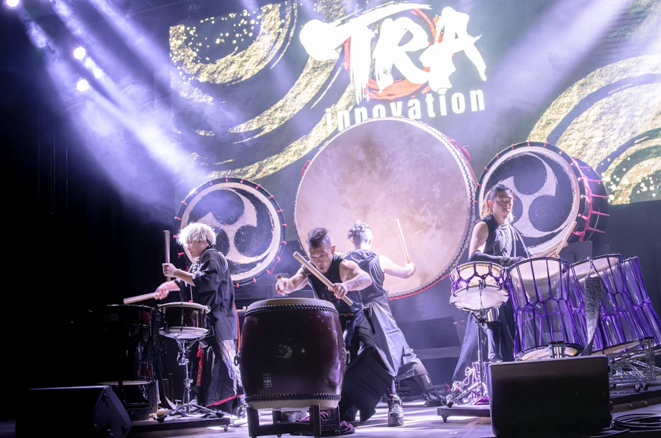 Four performers playing traditional drums on a stage with dramatic lighting and a banner reading "TRA Innovation."