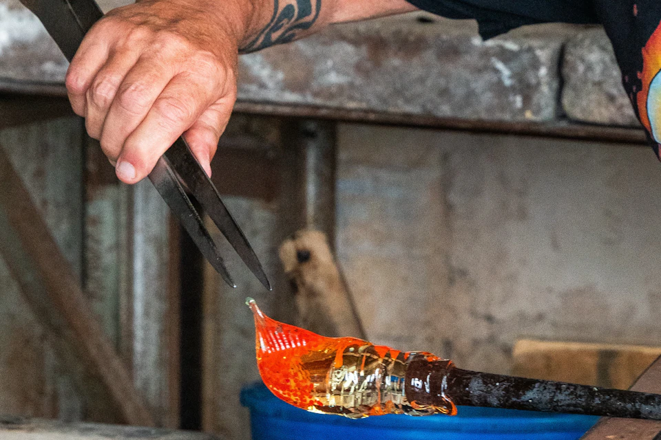 A person's tattooed arm uses tongs to shape molten glass, with equipment visible in the background.