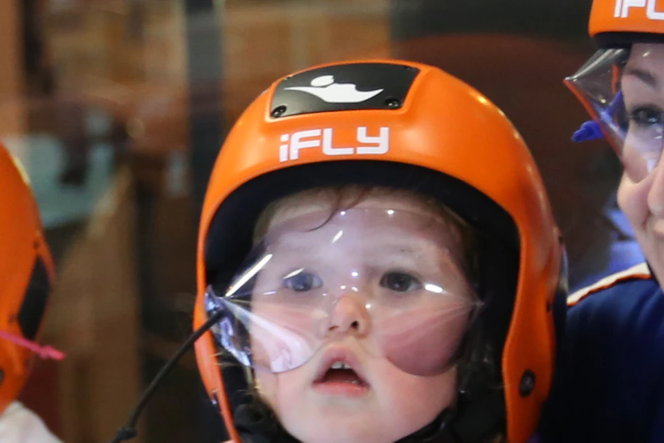 Adults and children in flight suits and helmets in an indoor skydiving facility, one adult holding a small child.