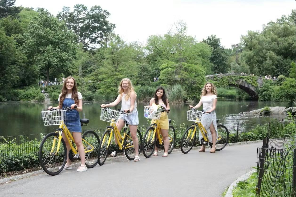 Three women standing next to yellow bicycles beside a pond in a park with lush greenery.