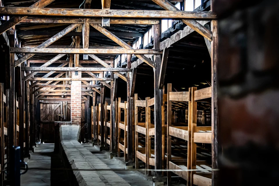A dimly lit, old wooden building interior with numerous wooden bunk beds and a pathway in the center.