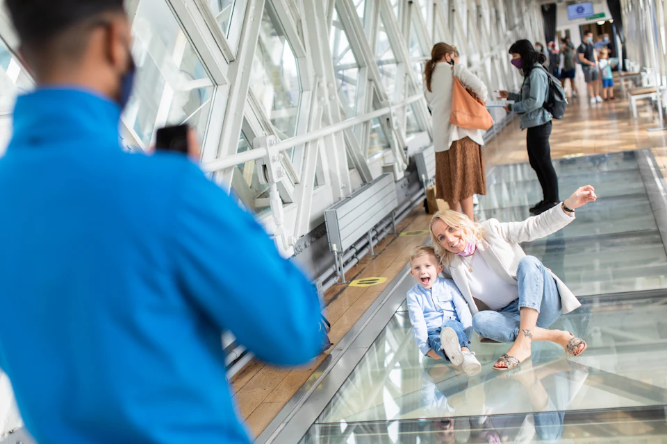 A woman and a child smile for a photo on a glass floor in a modern, indoor walkway. Other people walk and observe the area.
