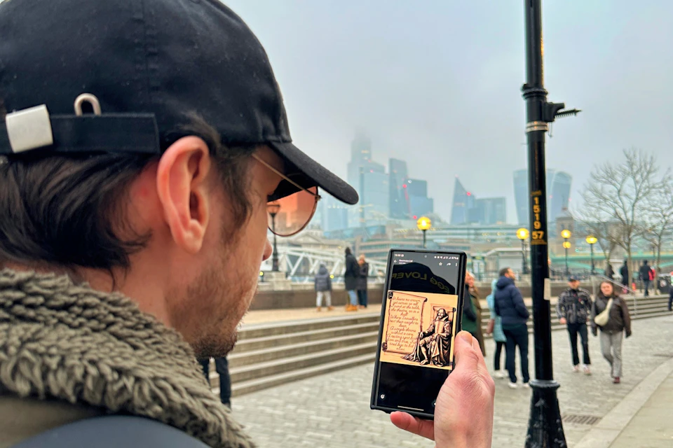 A person holding a phone displaying a digital artwork, with a cityscape and people walking in a foggy urban setting.