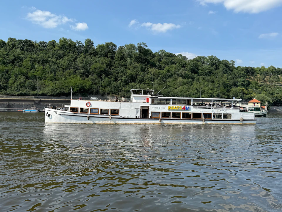 A white passenger boat on a river, with a forested shoreline in the background under a partly cloudy sky.