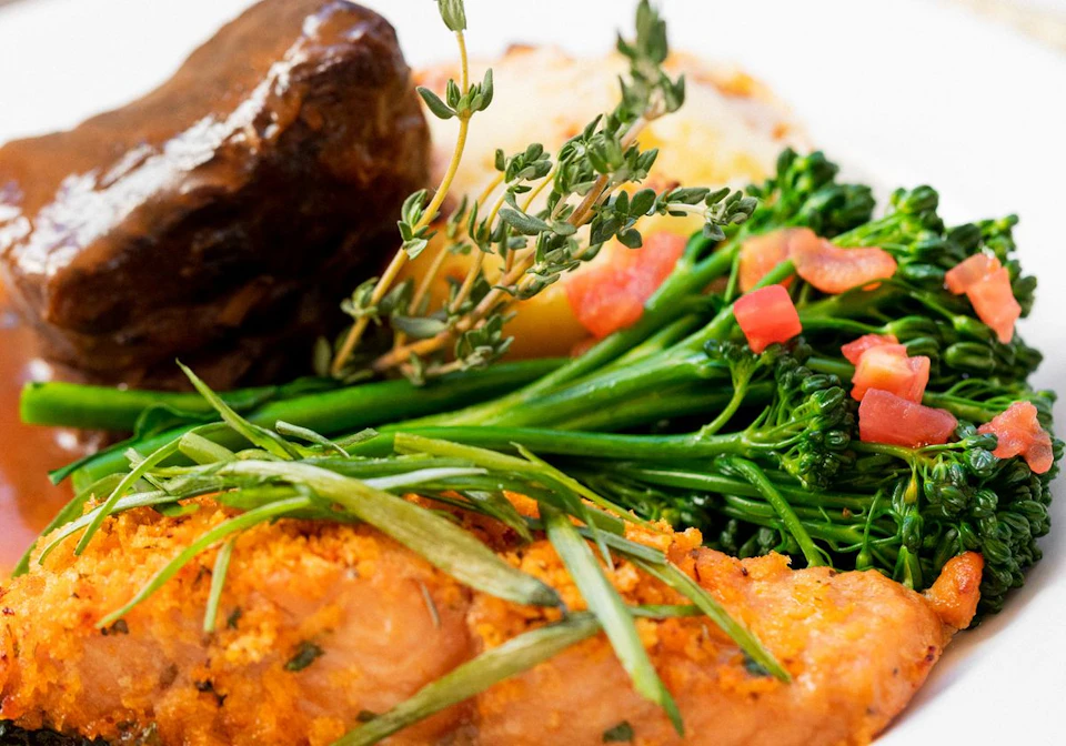 A close-up of a plated dish featuring roasted meat, broccolini, diced tomatoes, and herbs.