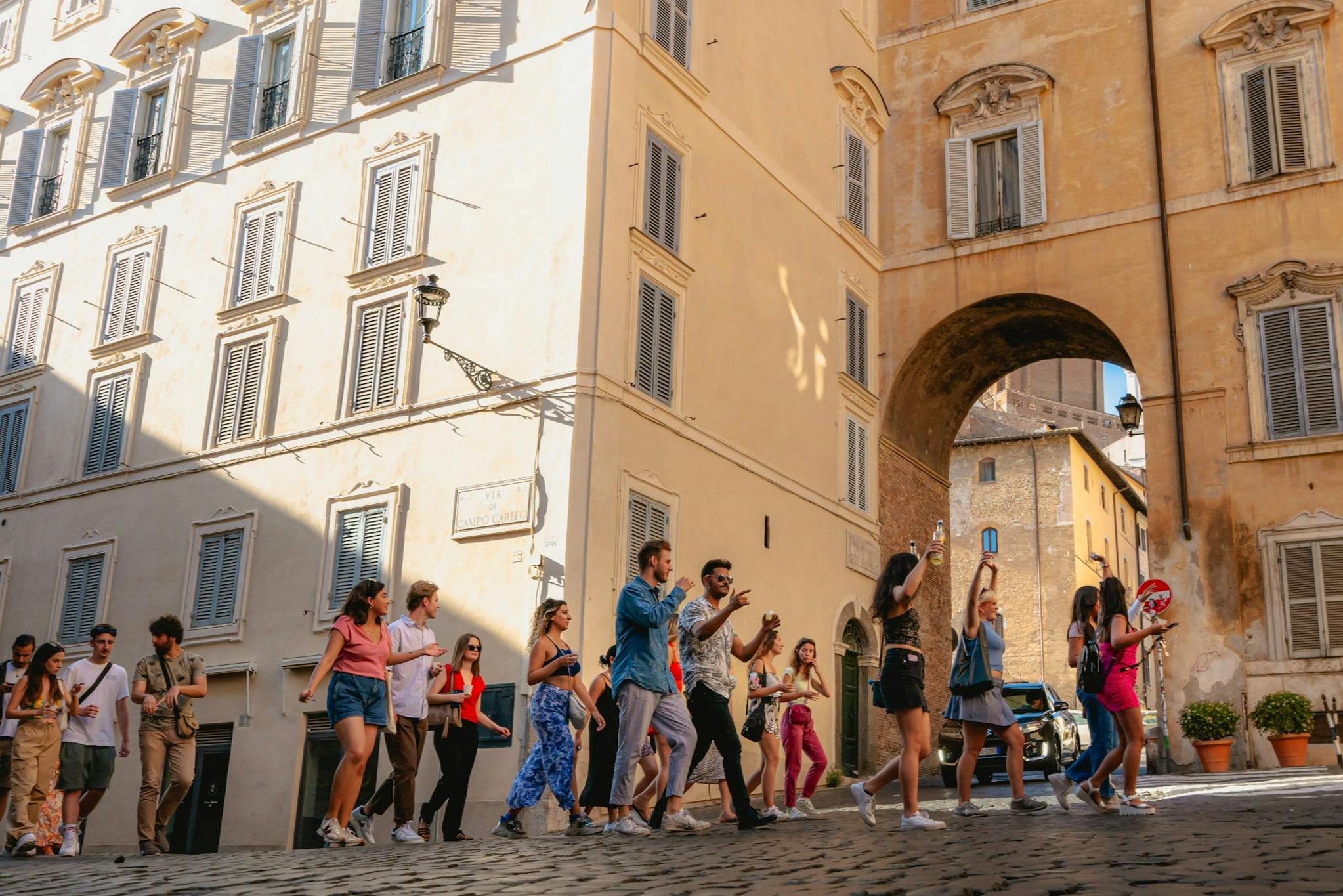 아치형 통로가 있는 유서 깊은 건물 옆 햇살 가득한 거리를 함께 걷고 있는 한 무리의 사람들.