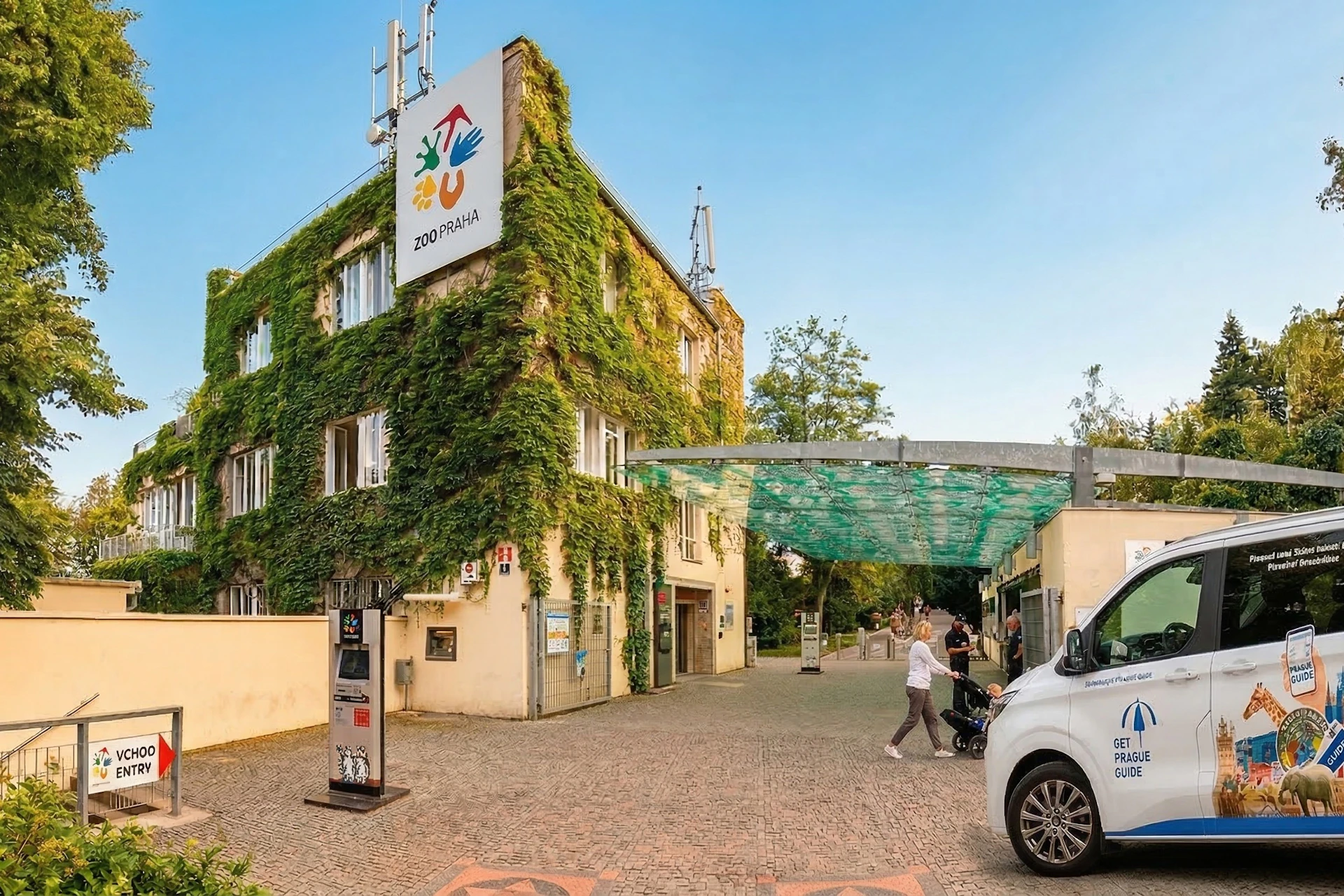 Entrance to Prague Zoo with a building covered in green ivy, a canopy structure, and visitors walking nearby.