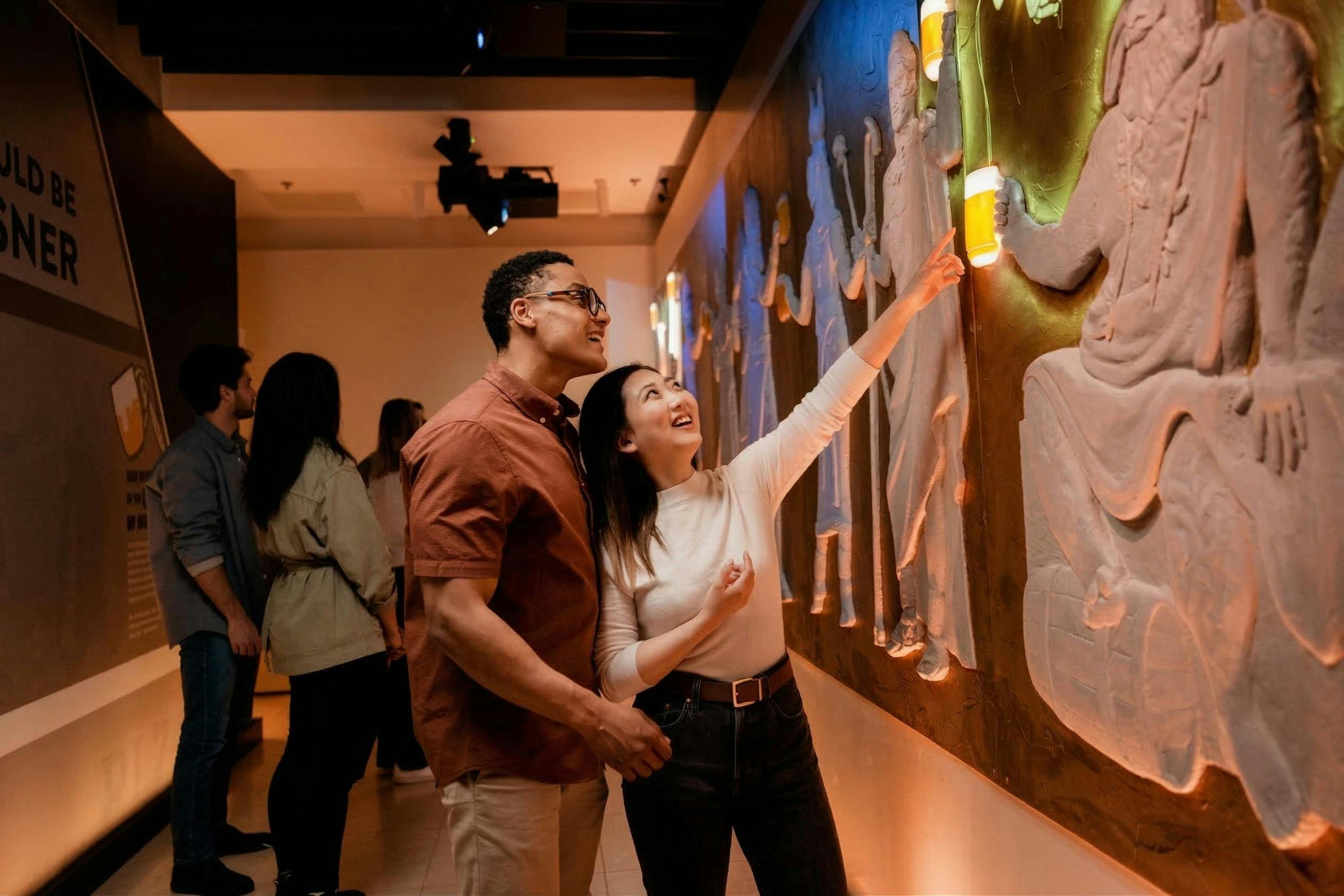 Ένα ζευγάρι παρατηρεί και αλληλεπιδρά με μια φωτισμένη έκθεση αρχαίων γλυπτών σε ένα μουσείο, με άλλους στο παρασκήνιο.