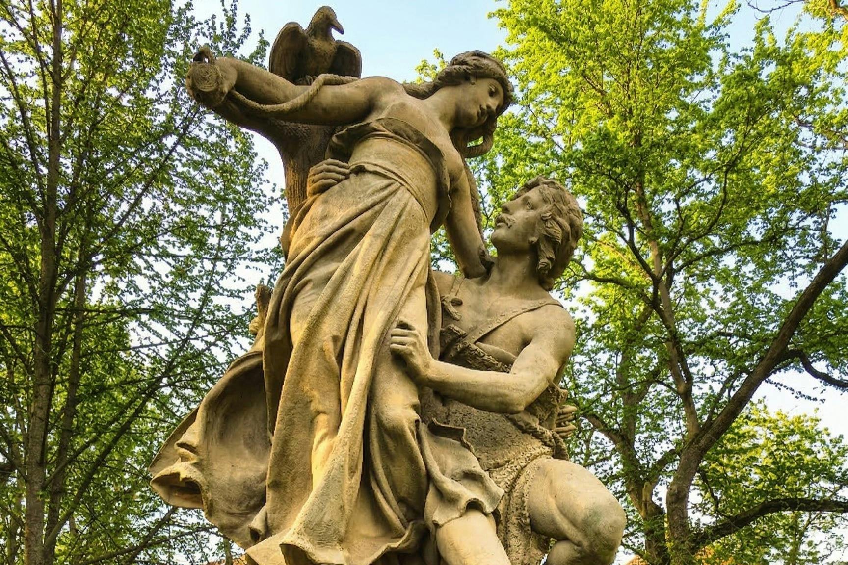 A classical-style statue of a man and a woman with an eagle, set against a background of green trees and blue sky.