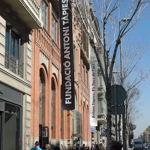 Människor går förbi Fundació Antoni Tàpies-byggnaden på en livlig gata kantad av träd och olika skyltfönster.