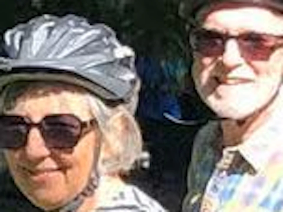 Two people wearing helmets pose with their bicycles on a park path surrounded by green grass and large trees on a sunny day.