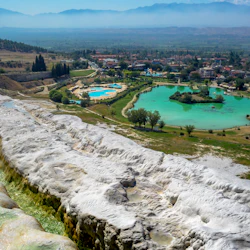 Terrasserade vita bergsformationer med grönt vatten; i fjärran finns en turkos damm, träd, byggnader och bergigt landskap.