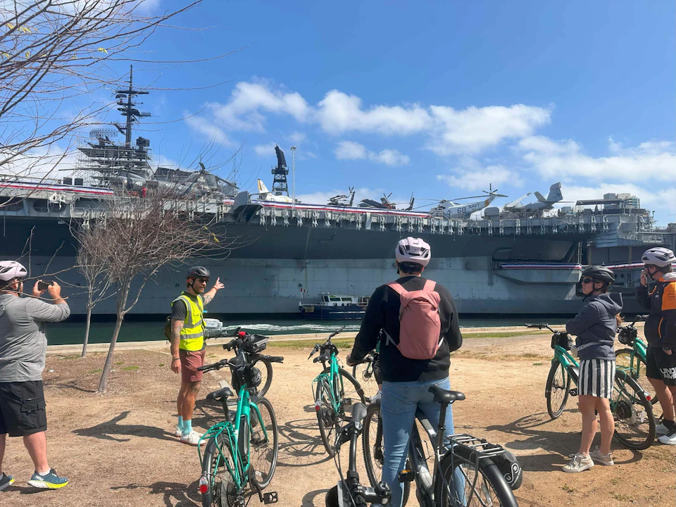 A group of cyclists views a docked aircraft carrier under a clear blue sky with scattered clouds.