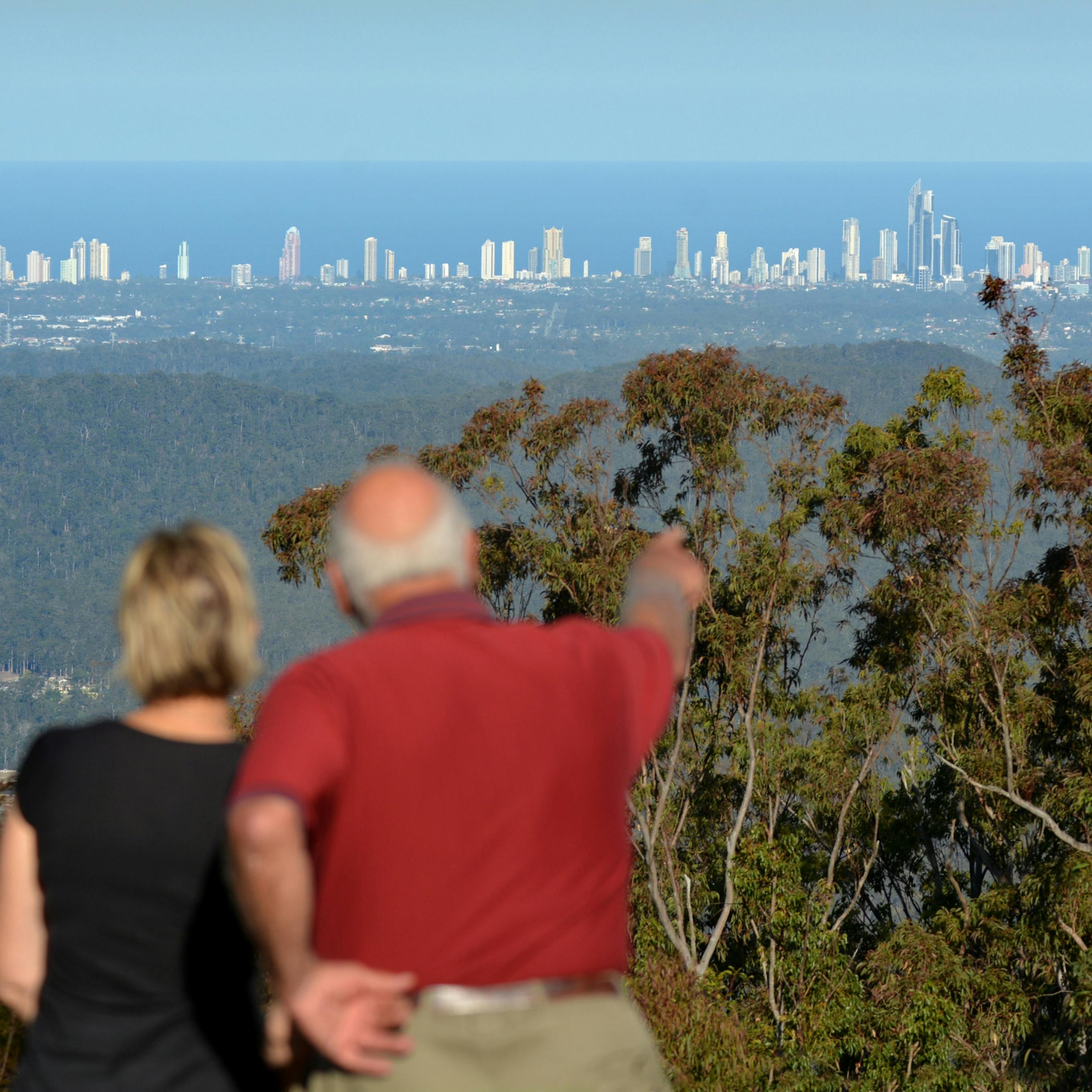 Deux personnes, dont l'une montre du doigt, observent la ligne d'horizon d'une ville lointaine avec de grands bâtiments, au-delà d'une zone boisée sous un ciel clair.